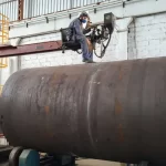 Operador soldando grande cilindro metálico com equipamento elevado.