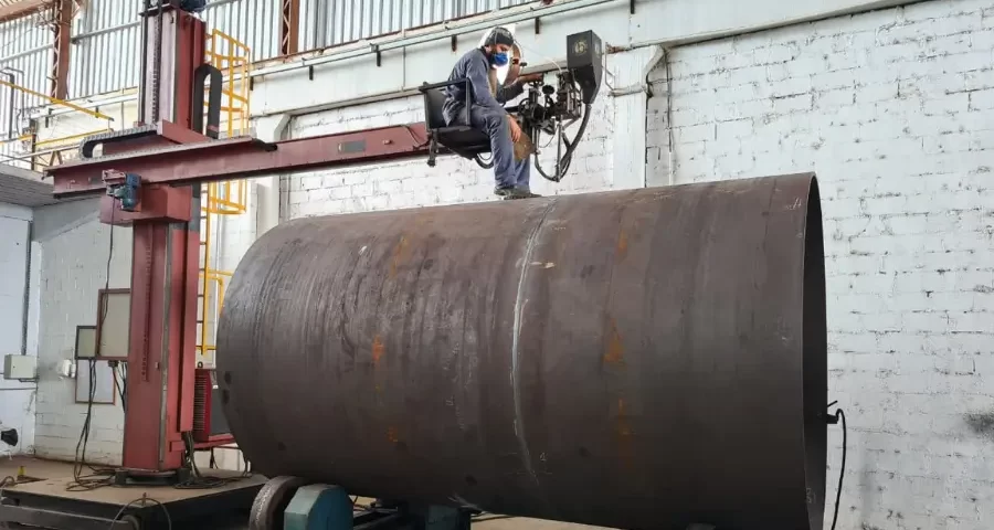 Operador soldando grande cilindro metálico com equipamento elevado.