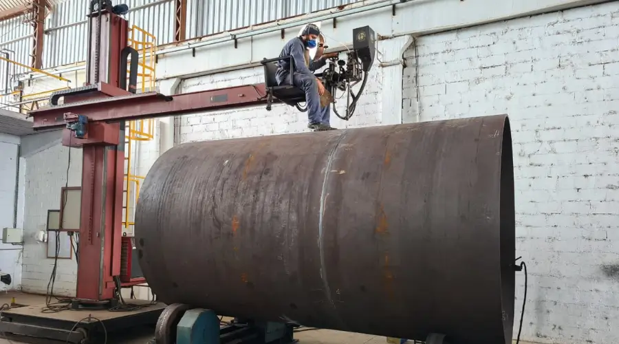 Operador soldando grande cilindro metálico com equipamento elevado.